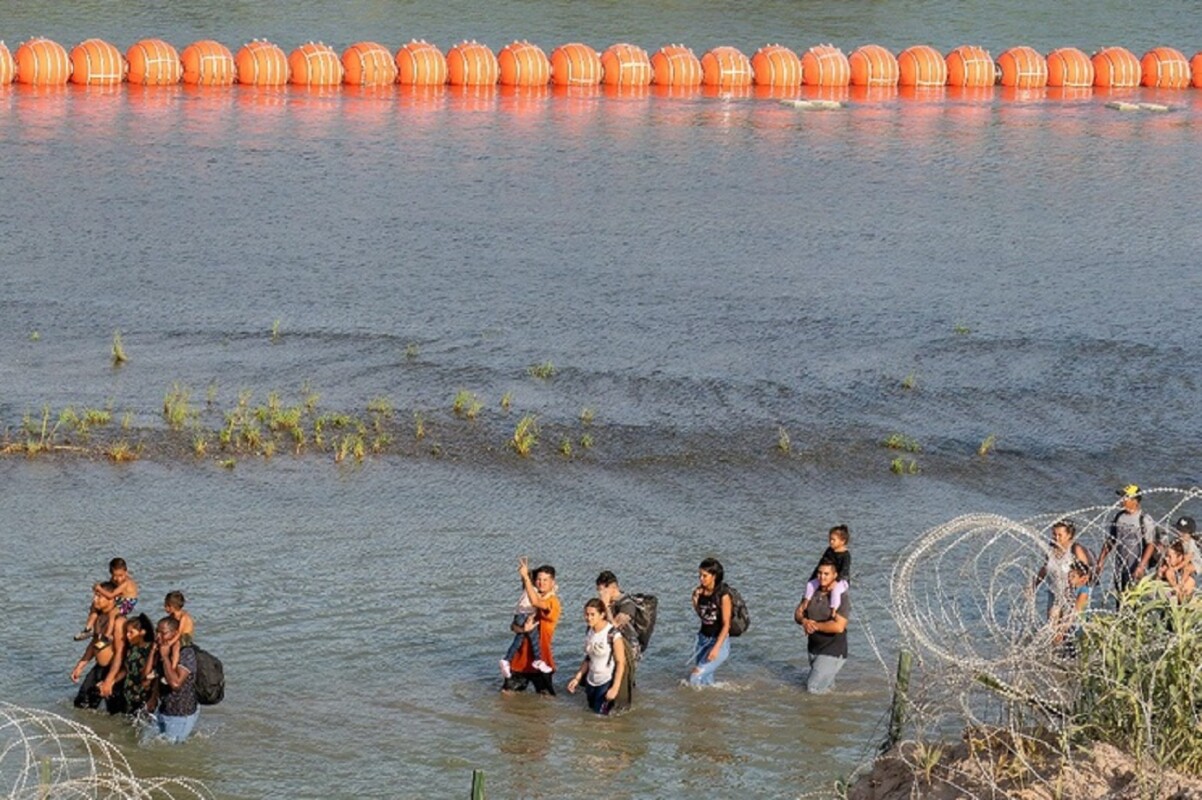 Migrantes caminan entre una valla de púas y una cadena de boyas en la línea divisoria con México en Eagle Pass, Texas. Foto Ap